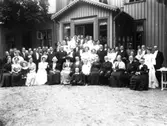 Karin Holmdahl, bröllop Tvååker. Gruppbild av brudpar med ett stort antal bröllopsgäster, fotograferade framför ett bostadshus. I bakre raden till höger står en yngling i uniform.