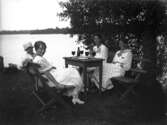 Fyra kvinnor sitter och äter vid trädgårdsmöbler vid sjöstrand. Ryd, Småland.