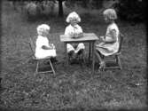 Tre småflickor sitter på barnstolar vid bord med leksak på i trådgård. Västerås.