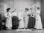 Sex utklädda personer. Figuren i vitt i mitten är en utklädd man, de övriga är damer. Maskerad eller ett teatersällskap.