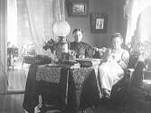 Bostadsinteriör där två kvinnor sitter vid ett bord, belamrat med stora böcker och en fotogenlampa. Kvinnan till höger ser ut att bära sjuksjöterskeuniform, så det kan vara en bostad relaterad till Varbergs lasarett. I ekononomibyggnaden från 1904 skulle det finnas bostäder åt operationssköterska, husmoder, tvättförestånderska och tvättpigor.