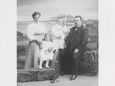 Familjebild av makar med två barn. Fond och rekvisita ska ge illusion av att de sitter på stenar vid havet, med Varbergs fästning i bakgrunden. Pojken håller i en åra.