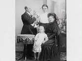 Familjebild med makar, två barn och en äldre svartklädd kvinna i stort dok. (Se även bild MR2_1016)
