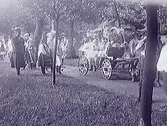 En sirligt dekorerad vagn dragen av häst på gångstig i parkliknande miljö. Vid sidan står åskådande barn, några i folkdräkter. Möjligen firande av Barnens Dag.