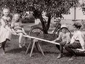 Två pojkar och två flickor på gungbräda i en trädgård. De är barn till föreståndarna för Mathilda Ranchs ateljé i Slöinge, Anna och Sven Christiansson. Fr v: Stina, Elna, Sven-Erik och Ivar. Vid trädet står en cykel.