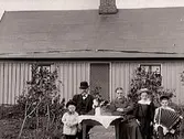 Man, kvinna och tre barn sitter vid ett bord framför ett trähus. Pojken spelar dragspel. Huset ser nybyggt ut och buskarna framför dörren är späda, så sannolikt är det en familj framför sitt hem.