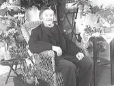 En herre som firar sin 90-åriga födelsedag sitter i en korgstol med gratulationsbuketter intill sig. Troligen en släkting till familjen Ranch. (Se även bild MR2_1313, 1315)