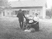 Kvinna och man med ett litet barn och en stor hund i trädgården med ett stort bostadshus i bakgrunden. Byggnaden har sticktak och stor veranda med snickarglädje.