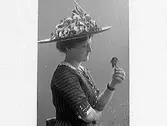 Kvinnoporträtt i profil. Fotografens syskonbarn, Bertha Larsson-Ranch, iklädd en stor hatt. Hon håller upp en blomma och tittar på den.