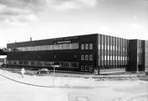 Gatuvy som visar industribyggnaden Sieverts kabelverk, korsningen Hemvägen/Lärkvägen i kv. Linet. Framför byggnaden en personbil och en kvinna.