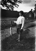 Barfota pojke med gädda på kroken, troligen Evert Klasson i Albäck. I bakgrunden syns en stuga med grödor på åkern längs vägen.