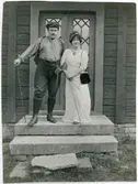 Olof Thunman med ryggsäck och vandringsstav står med en kvinna framför en stuga, Ängarås, Västergötland 1909