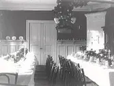 Uppdukade långbord i en sal med kakelugn och dubbeldörrar. Takarmaturen är lövad.