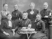 Gruppbild av sju kvinnor. Eventuellt en föreningsgrupp av något slag, eller en personalgrupp. På bordet står ett fotografi av ytterligare en person. Elisabet Bolin är beställare.