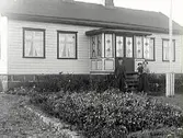 Exteriör av villa med glasveranda och liggande träpanel. Ett par står med litet barn framför verandan. Vid flaggstången en rabatt eller möjligen potatisland.