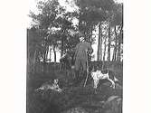 En jägare med keps och bössa står i skogen med två hundar och håller upp en död räv. (Se även bild MR2_1598)