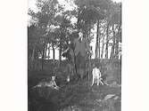 En jägare med hatt och bössa står i skogen med två hundar och håller upp en död räv. (Se även bild MR2_1597)