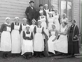Troligen en personalgrupp med två män och övriga kvinnor, vilka alla bär förkläden utom en äldre kvinna klädd i klänning och kofta. Troligen köks- eller vårdpersonal av något slag. De står samlade kring en byggnads trappa utomhus.