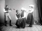 Folkdans. En fiolspelare, en kvinna som sitter i knät på en man och till höger om dem en kvinna som håller ut händerna mot axlarna på flickan i knät. (Samma fiolspelare på bilderna MR2_1699, 1735, 1938)