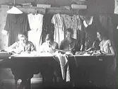 Sömmerskor. Ester och Jenny Nilssons syateljé i Stålbåges hus, Prästgatan i Varberg. Jenny står vid bordet där sömmerskorna sitter och bakom dem hänger klänningar på rad.
Från höger: Elin Andersson från Bua, Ebba från Horred, Ester Nilsson från Varberg, Jenny Nilsson från Varberg, Inghammar från Sibbarp-Dagsås.