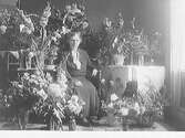 En äldre kvinna, fru Bengtsson, Pensionatet, sitter omgiven av ett stort antal blomsterbuketter. Födelsedagsfirande.