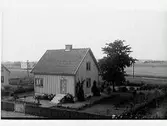 Exteriör av bostadshus och trädgård tillhörigt en herr Ljung i Träslöv. I bakgrunden ett jordbrukslandskap.
