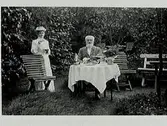Musikdirektör Alfred Lundgren med hushållerska Anna Bäckström från Värmland, fotograferade i trädgården på fastigheten Klockaren 18, Varberg. Alfred sitter vid det dukade kaffebordet medan Anna står bredvid med koppen i hand.