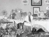 Apotekare Hultgrens fru Jenny firar födelsedag. hon sitter i salongen omgiven av blomster. Hennes sjal har liksom bordsduken långa fransar. Bostaden ligger i kv Apotekaren 8, hörnet av Torggatan-Drottninggatan.