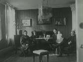 Familjebild hemma hos Adolf Andersson. Tre vuxna och två barn sitter i salongens soffgrupp, där kakelugnen skymtar i höger hörn. Den tidstypiska jugendarmaturen i taket är märkligt nog inplastad.