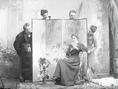 Lek i ateljén kring en vikskärm. Bakom skärmen från vänster: fru Hedvig Zetterberg, Elof Ernwald, Hulda Lindh, Wilhelm Ranch, Mathilda Ranch. Längst fram sitter fröken Gustavsson med en bok.