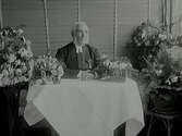 Prosten Magnus Ullmans 80-års dag. Han sitter vid ett bord med duk, omgiven av gratulationsblomster. Bakom honom hänger träjalusier.