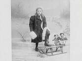Ateljébild av fotografens systerdotter. Hon är vinterklädd mot snöfond och har ena foten på en kälke, där det sitter leksaker baktill.