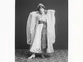 Ateljéfoto av kvinna i speciell klädsel med slöjor över armarna och brett textilskärp lika långt som klänningen. Kanske en aktris. (Se även MR2_929, MR2_950 och MR2_969)