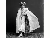 Ateljébild av kvinna i speciell klädsel med beslöjade armar och brett, långt textilskärp med fransar. Kanske någon sorts aktris eller utländsk klädsel. (Se även MR2_ 931, MR2_950 och MR2_969, MR2_975, MR2_976)