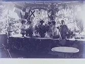 Veranda i park/trädgård med ett långt bord med blombuketter i förgrunden och folk i bakgrunden. Ett flaggspel hänger i taket. Troligen från en utställning i societetsparken.