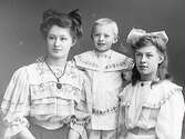 Ateljébild av tre syskon, troligen. En liten pojke och två flickor. Pojken bär smalrandig, broderad kolt med krage som täcker axlarna.