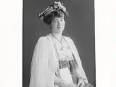 Ateljéporträtt av exklusivt klädd kvinna med slöjor över armarna och ett brett, textilt skärp. Platt, blomstersmyckad hatt. Möjligen utsländsk klädsel. Om halsen hänger en kuvertliknande väska.
(Se även bilderna MR2_931, 950, 969, 976)