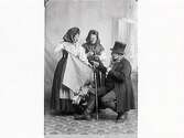Tablå med Västgötaknalle i hög hatt och käpp/stav som sittande säljer tyg till två stående kvinnor i hucklen. Ena damen är Hulda Lindh (f Ranch) och mannen är Elof Ernwald. (Jämför bild MR2_985 o 983)