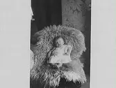 Barnporträtt. Baby som ligger i en stol med fårfäll. I bakgrunden en tapet med mönster av stora kastanjeblad.