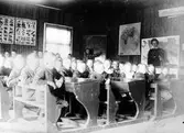 Agnsjöns skola, Hemsjö socken. Interiör från skolsalen. En lärarinna i katedern och elever som sitter i skolbänkarna. Originalet på dagsljuspapper i vykortsformat.