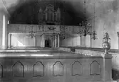 Interiör av okänd kyrka. Med altaret bakom oss ser vi kyrkbänkar, ljuskronor och orgeln uppe på orgelläktaren.