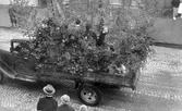 Barnens dag 1938. Lövat lastbilsflak med kvinnor och män, passerar på gatan där en folksamling står.