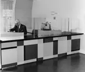 1955 års postkontor, rum nr 210 på 2 trappor. Vid den halvfrontala
disken i kassaexpeditionen agerar herr Votele Org. (I fonden fönster
mot Munkbrogatan)