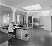 1955 års postkontor, rum nr 210 på 2 trappor. Vid den halvfrontala
disken i kassaexpeditionen agerar överpostexpeditör Majken
Cederberg-Olsson museibesökare. (Till vänster fönster mot gården, i
fonden öppning till rum 212).