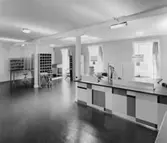 1955 års postkontor, rum nr 210 på 2 trappor. Vid den halvfrontala
disken i kassaexpeditionen agerar överpostexpeditör Majken
Cederberg-Olsson museibesökare. (Fönster mot Munkbrogatan)