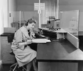 1955 års postkontor, rum nr 210 på 2 trappor. Vid den halvfrontala
disken i kassaexpeditionen agerar extra ordinarie kontorsbiträdet
Birgitta A.L. Nyberg museibesökare. (I fonden fönster åt gården).