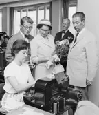 Besök på bokföringsavdelning E. Kontorsbiträder fru Görel Remling
stansar hålkort för bokföring på herr Malmréns konto. Tf girobokföringschefen överkontrollör Gillis Schreiber t.h. övervakar.
Postsparbanksintendenten E.G. Carlsson i bakgrunden. Fru Inga Malmrén har blivit uppvaktad med blommor av girokontoret.