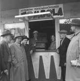 En grupp människor har samlats utanför Huskvarna pensionärsförenings Tombola lotteri, på Esplanaden i Huskvarna året är 1957.