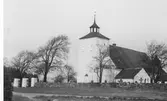Onsala kyrka och kyrkogård sedd från sydväst. Mitt för tornet ses entrépartiet som tillkom 1836 med dubbla grindar mellan tre höga, fyrkantiga pelare. Kyrkogården hägnas av en stengärdsgård.