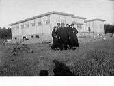 Gruppbilder av personer framför byggnaden på en barnkoloni i Gamla Köpstad. I mitten av gruppen står förmodligen Sven Larsson, ägaren till marken. Runt honom troligen medlemmar ur Ludvika Barnens dagskommittén.
